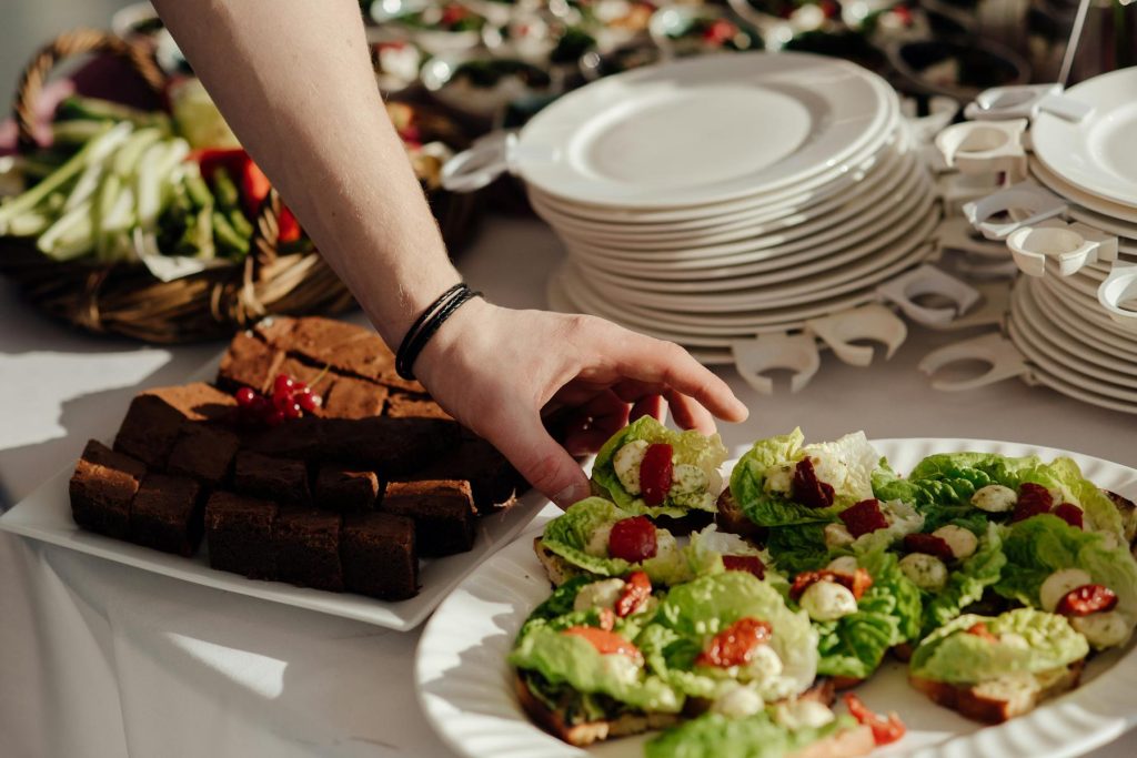 Crop anonymous man taking snack with lettuce and cherry tomatoes with cheese from buffet table with brownie during banquet in restaurant